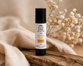 A lip balm tube labeled 'Hearth & Hide Golden Peaks Lip Balm' standing upright on a rustic wooden surface, surrounded by soft beige fabric and dried white flowers in a warm, softly lit setting.