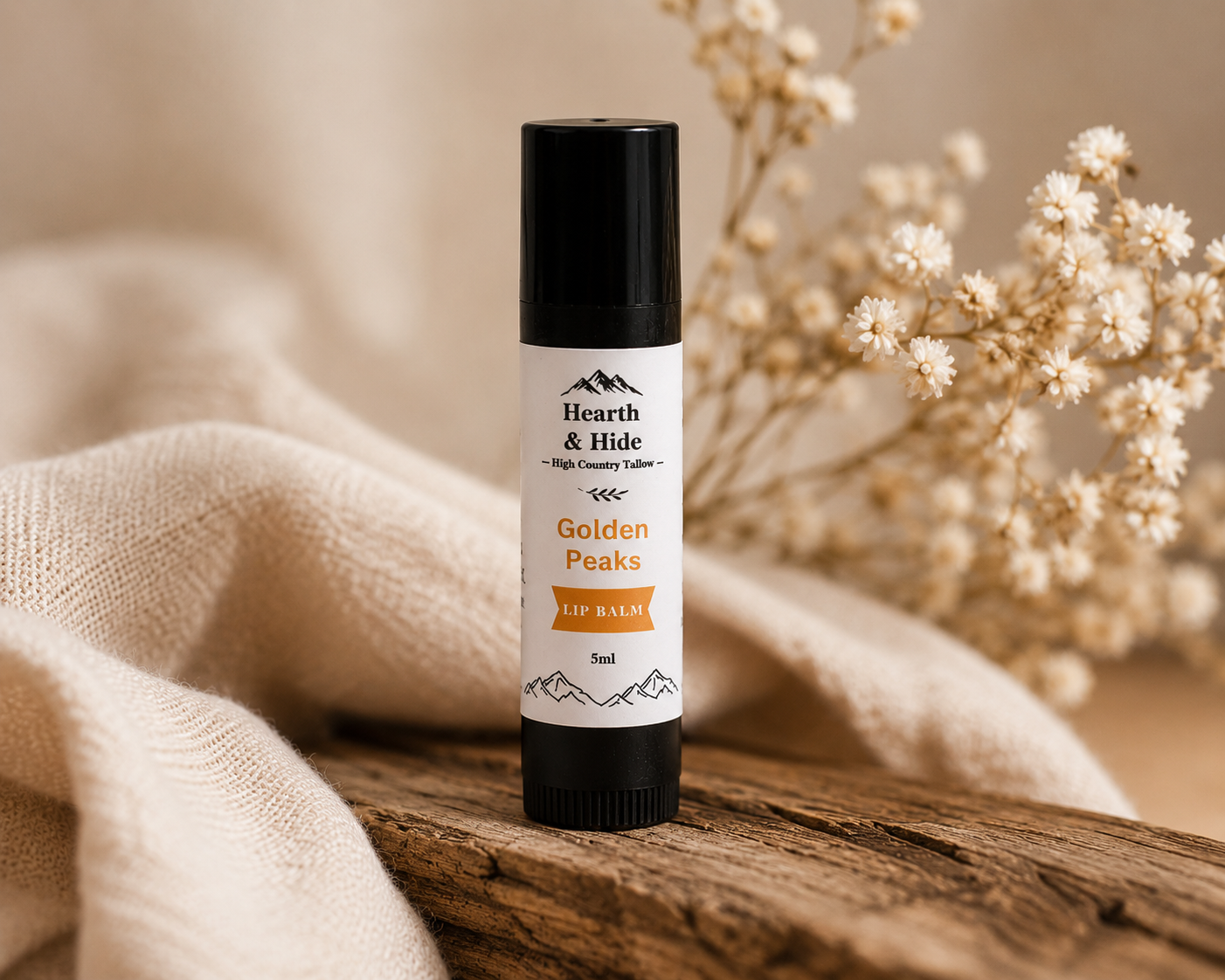 A lip balm tube labeled 'Hearth & Hide Golden Peaks Lip Balm' standing upright on a rustic wooden surface, surrounded by soft beige fabric and dried white flowers in a warm, softly lit setting.
