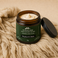 An open amber glass jar of Shepherd's tallow Balm with a dark green label and black lid, resting on a chunky beige knit blanket.