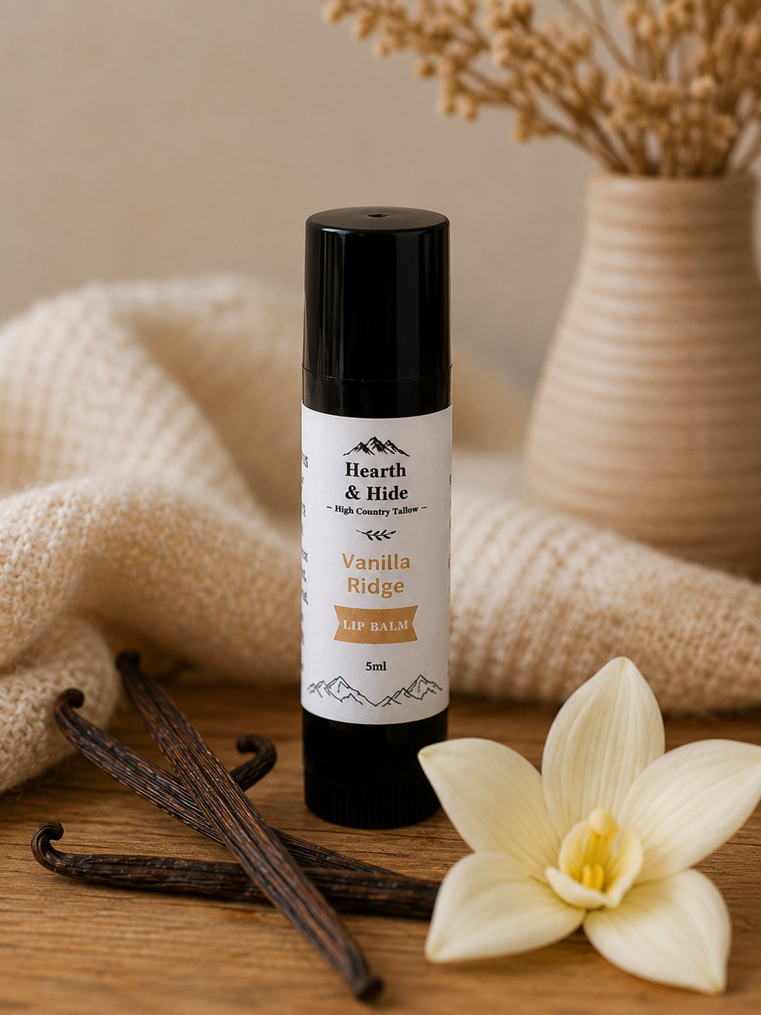 A 5ml tube of Hearth & Hide Vanilla Ridge Lip Balm stands on a wooden surface, styled with vanilla beans and a white vanilla flower, with a cozy beige knit blanket and dried flowers in the background.