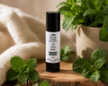 A tube of lip balm standing upright on a wooden surface, surrounded by fresh mint leaves and a soft beige fabric backdrop. The label reads 'Hearth & Hide' and 'Wild Mint Lip Balm.