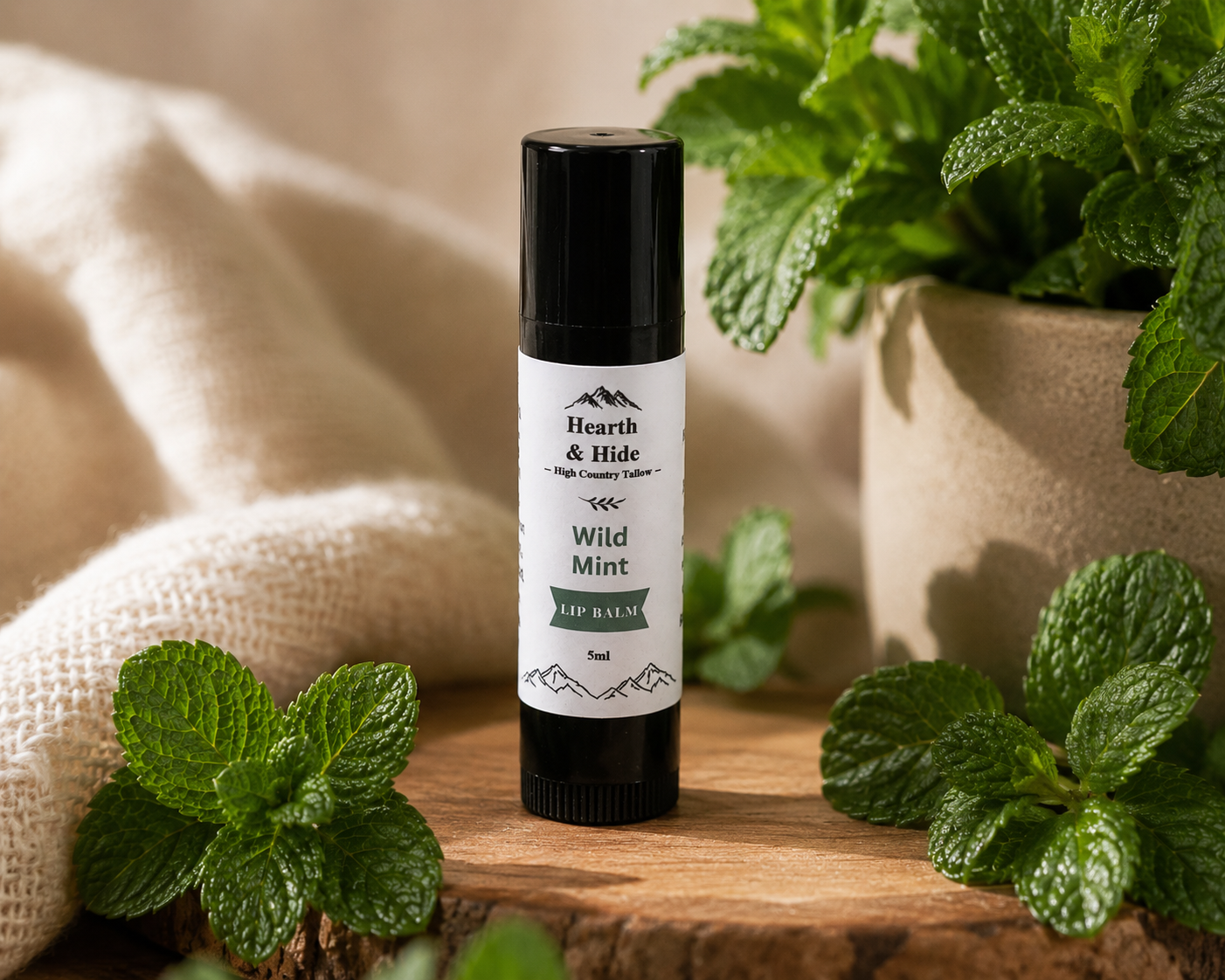 A tube of lip balm standing upright on a wooden surface, surrounded by fresh mint leaves and a soft beige fabric backdrop. The label reads 'Hearth & Hide' and 'Wild Mint Lip Balm.
